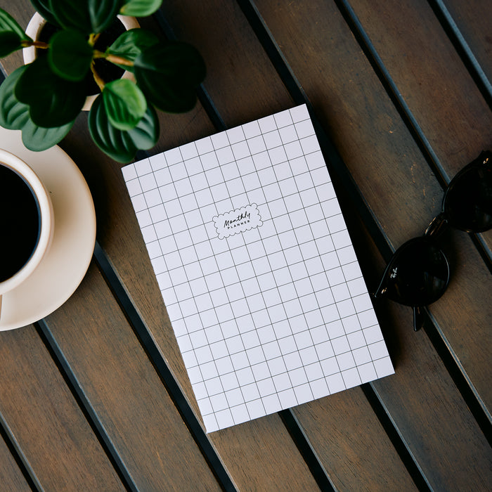 Undated Modern Tones Folio Planner Insert - Monthly Calendar View A5 Layout- 12 Months shown as a white grid planner page on a wooden table with coffee and greenery beside it