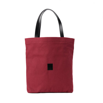 Red canvas shoulder bag with black handles and a small front patch on white background