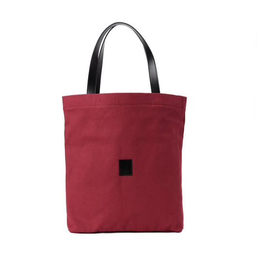 Red canvas shoulder bag with black handles and a small front patch on white background