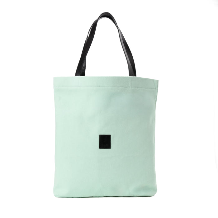Light green canvas tote bag with black handles and a black square patch on front