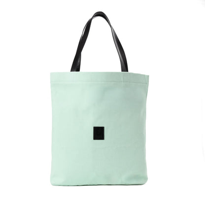 Light green canvas tote bag with black handles and a black square patch on front