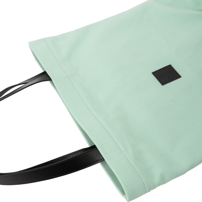 Mint green canvas tote bag with black leather handles and a black square patch
