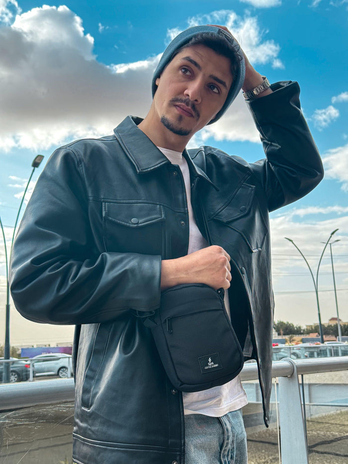 Man in black leather jacket and beanie wearing black cross bag outdoors